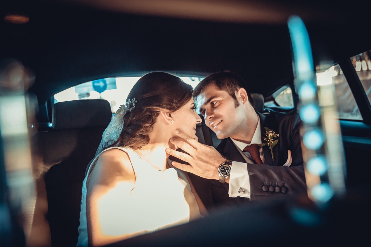 Pareja de novios besándose en el interior de su coche de bodas, acariciándose. Con rayos de luz que entran por las ventanillas